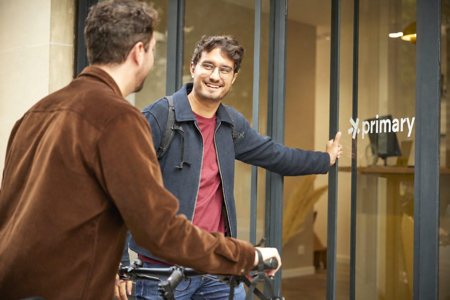 Deux hommes se saluent devant l'entrée vitrée d'un cabinet Primary, l'un tenant un vélo, le logo Primary visible sur la vitre.